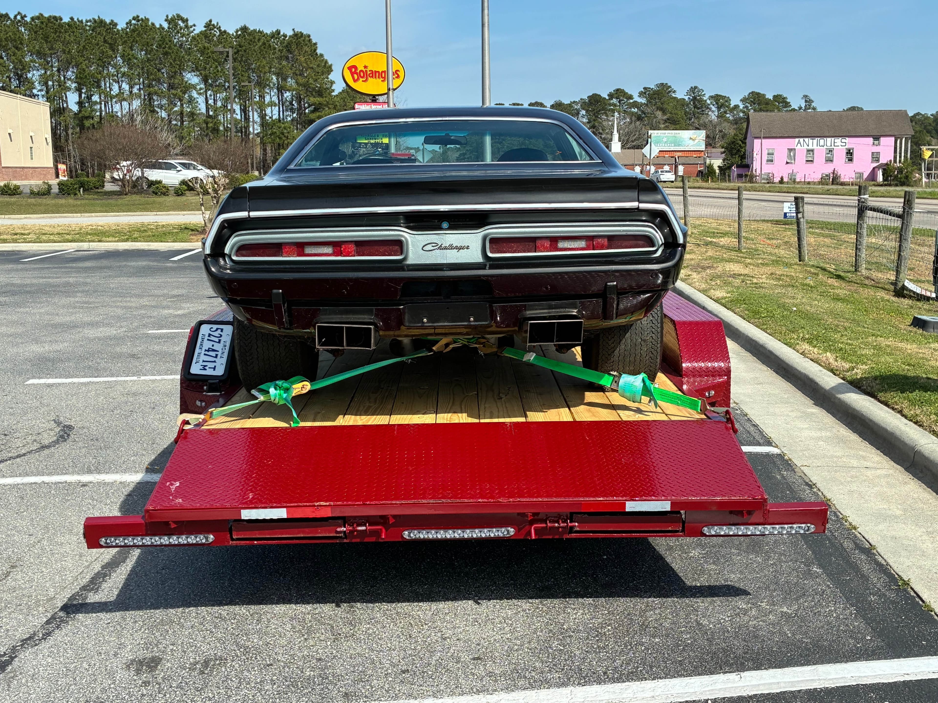 Black and dark green 1970s Dodge Challenger muscle car on red cargo trailer with green tie-down straps, rear view