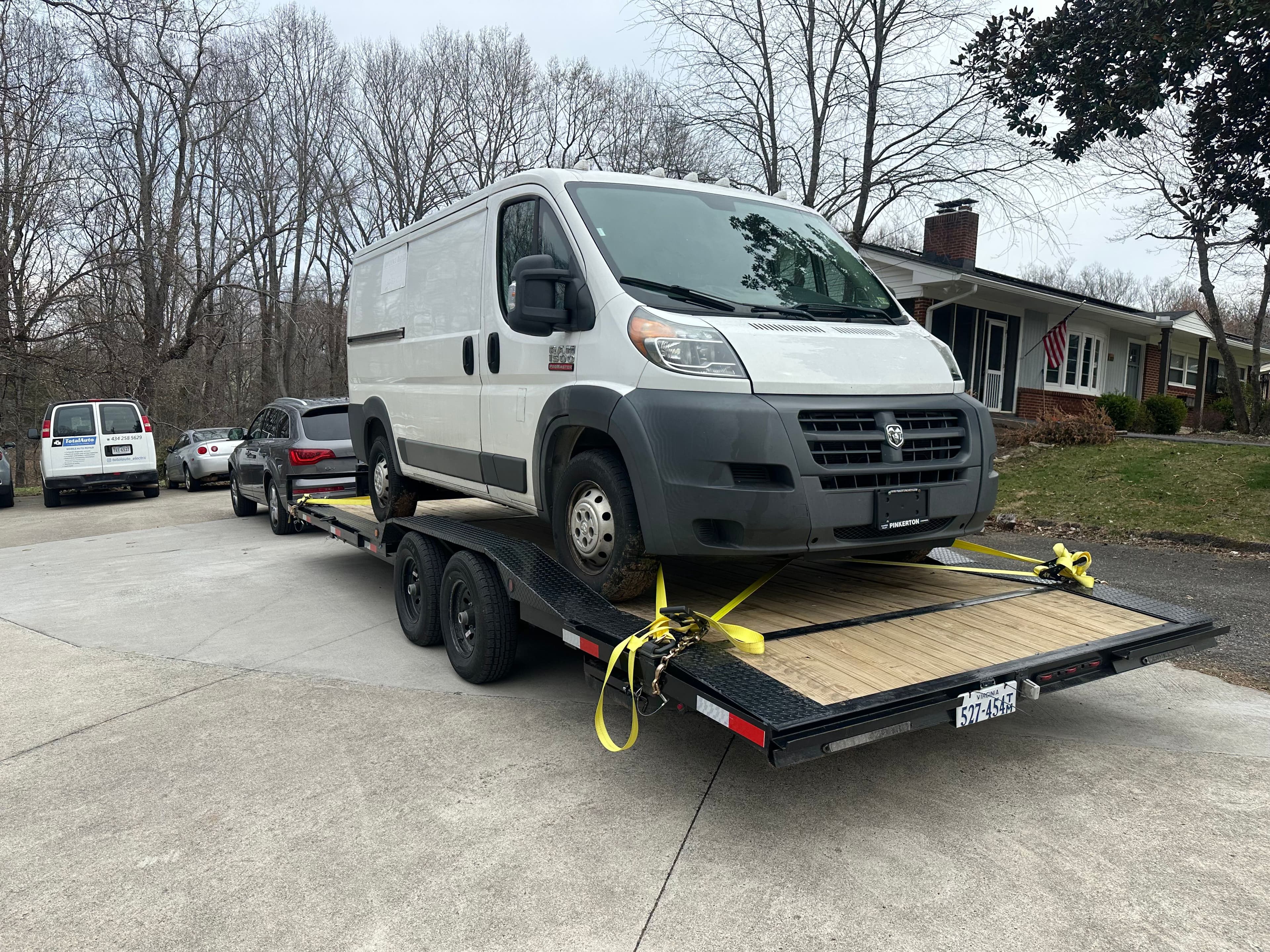 White work van on black dual-axle car trailer with yellow tie-down straps in residential driveway setting