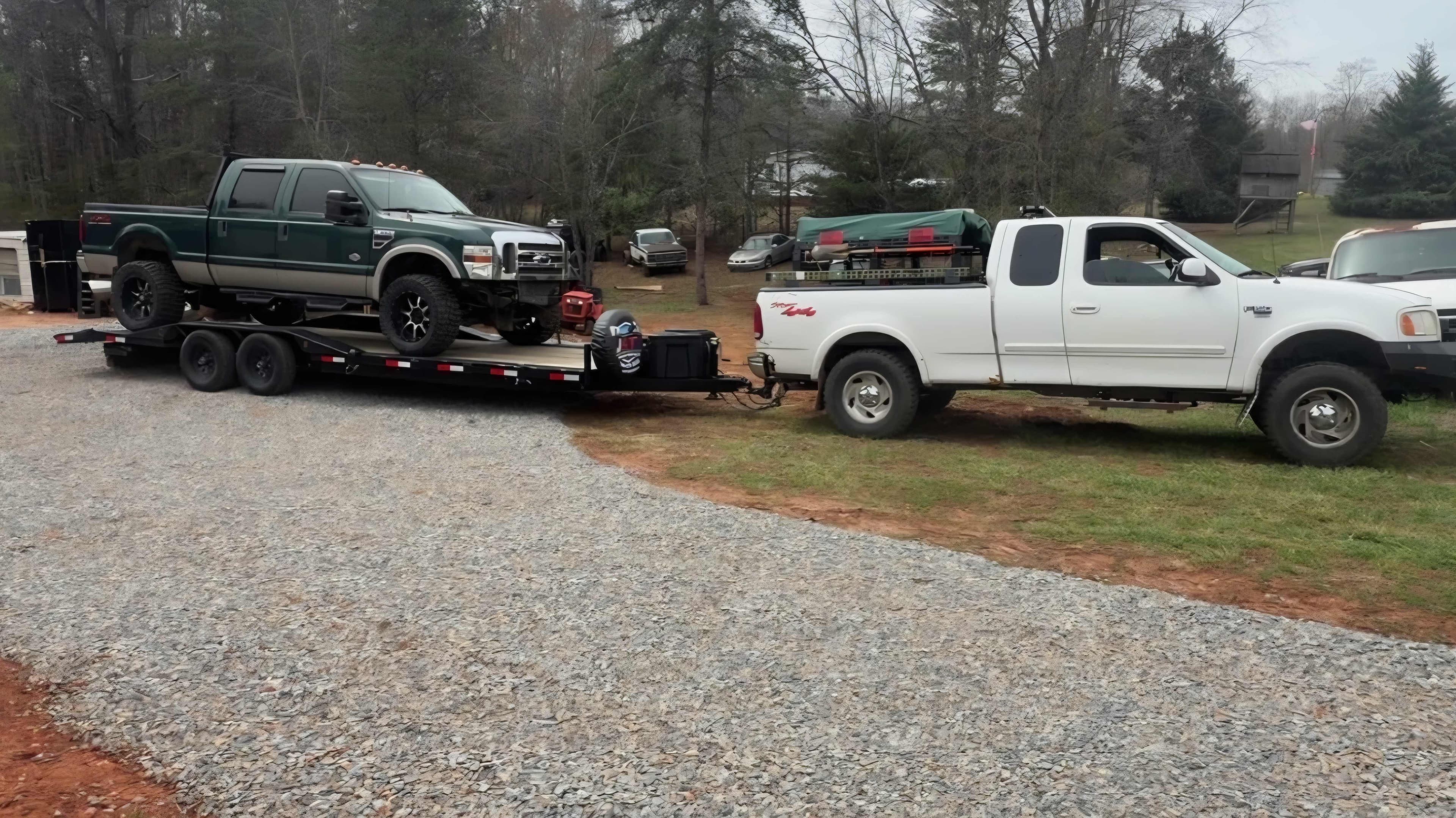 White Ford F-150 towing a black dual-axle trailer loaded with a lifted green Ford F-250 Super Duty truck on a rural gravel driveway