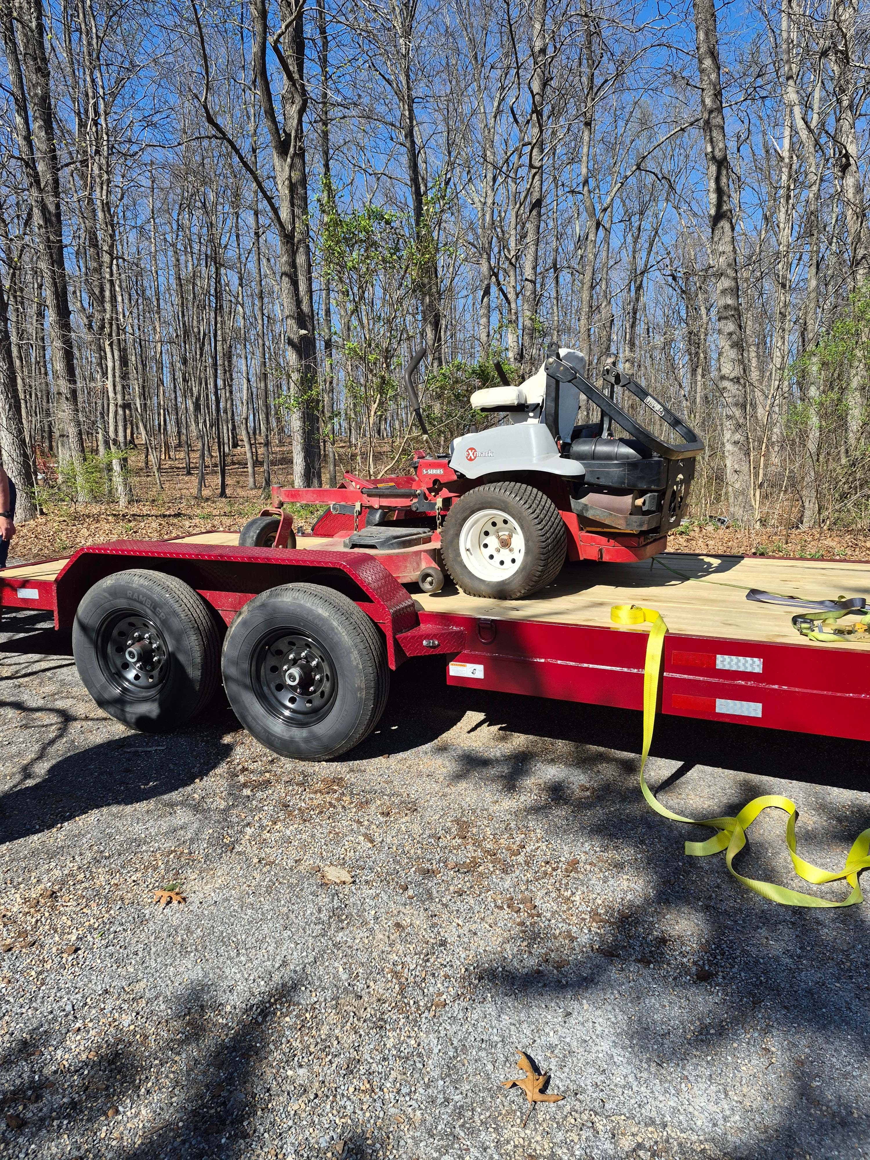 Exmark S-Series zero-turn riding lawn mower loaded on a red dual-axle cargo trailer with yellow tie-down straps on a gravel driveway