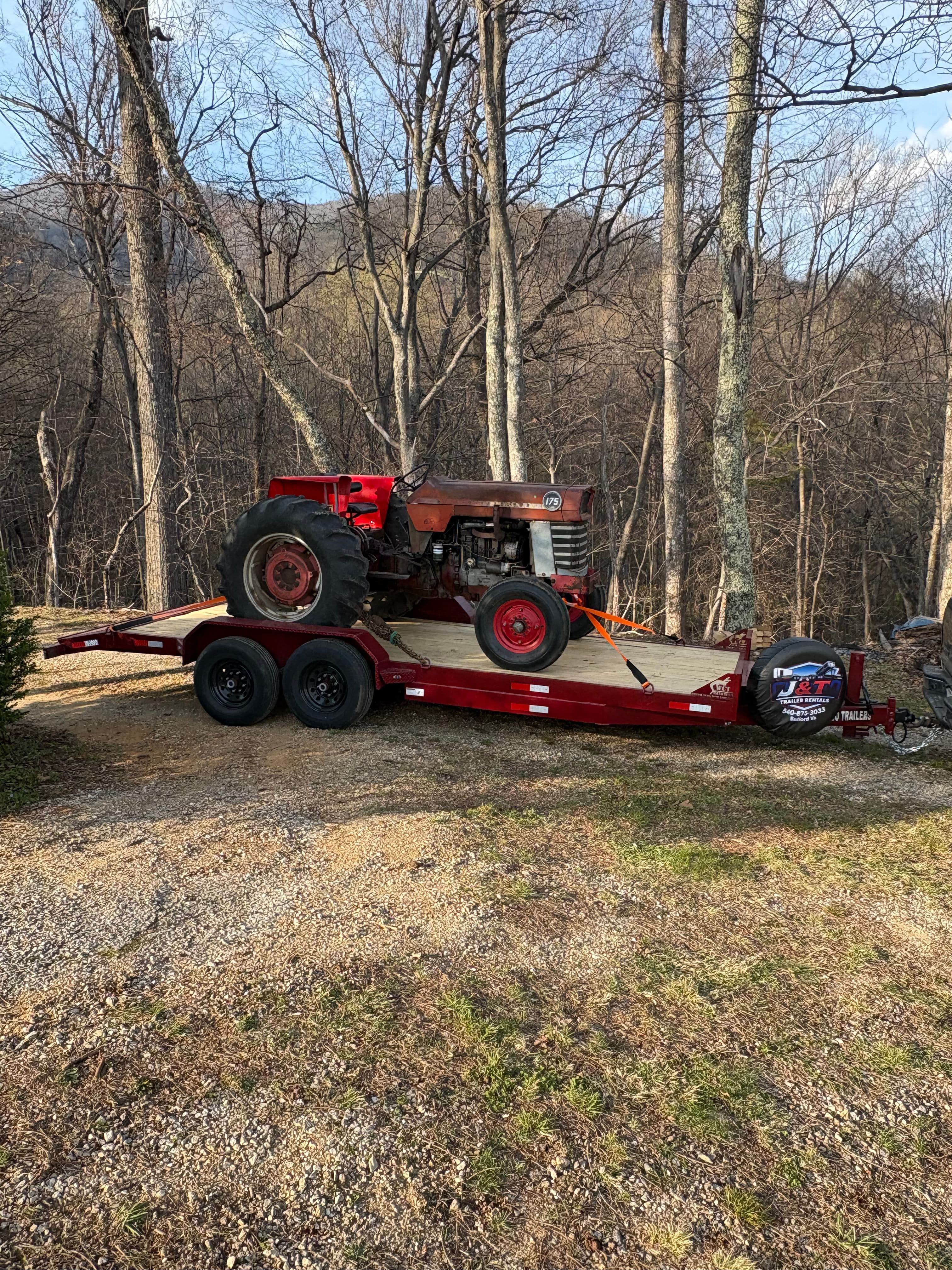 Massey Ferguson 175 vintage tractor on a J&T Trailer Rentals red dual-axle cargo trailer, full profile view in a wooded gravel driveway