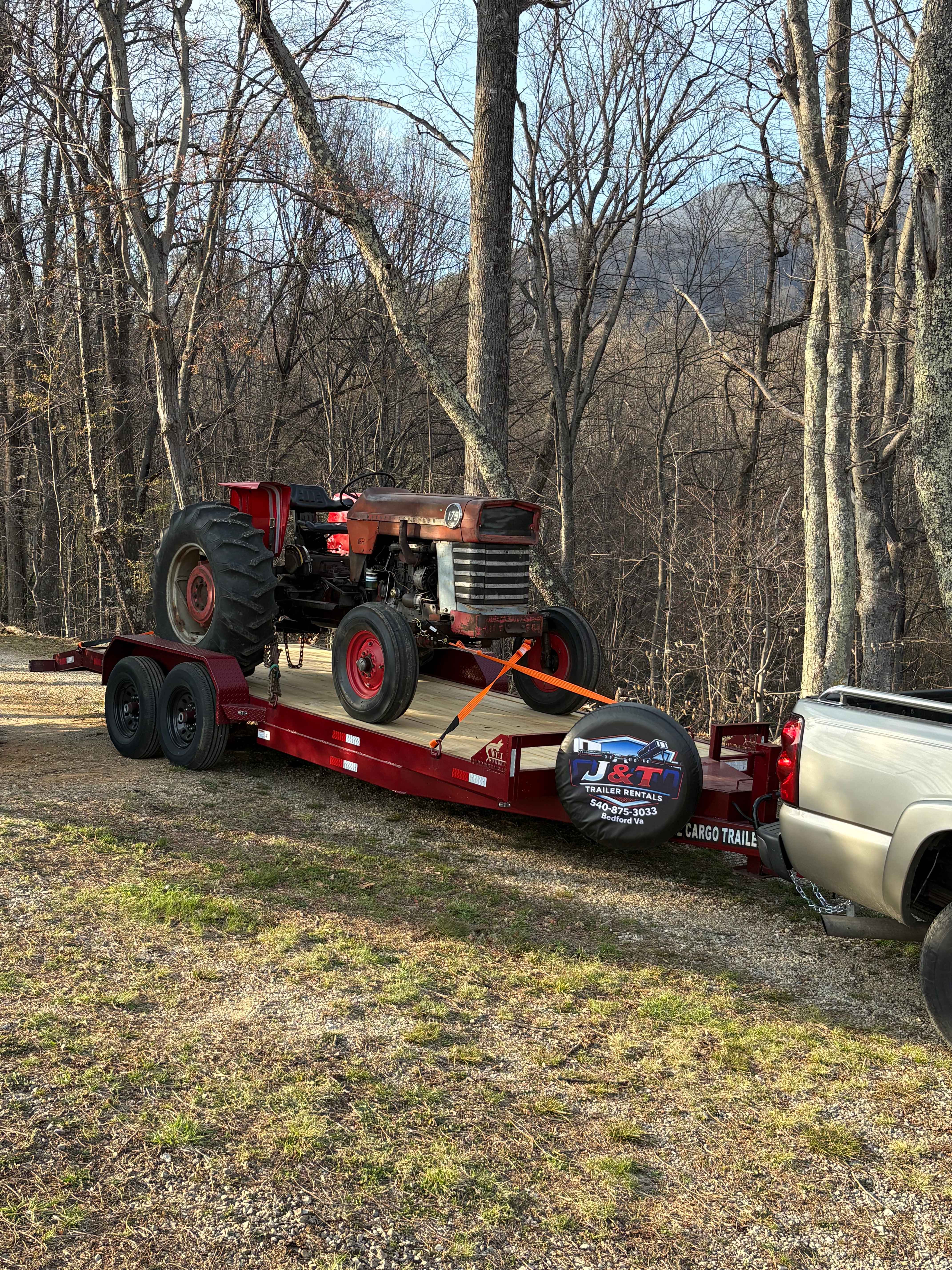 Vintage red farm tractor secured with orange tie-down straps on a J&T Trailer Rentals red dual-axle cargo trailer in a wooded rural setting