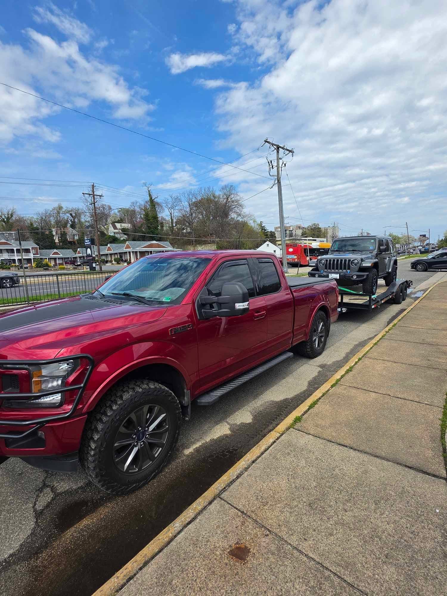 Red lifted Ford F-150 towing a black dual-axle car trailer with a gray Jeep Wrangler loaded on an urban street