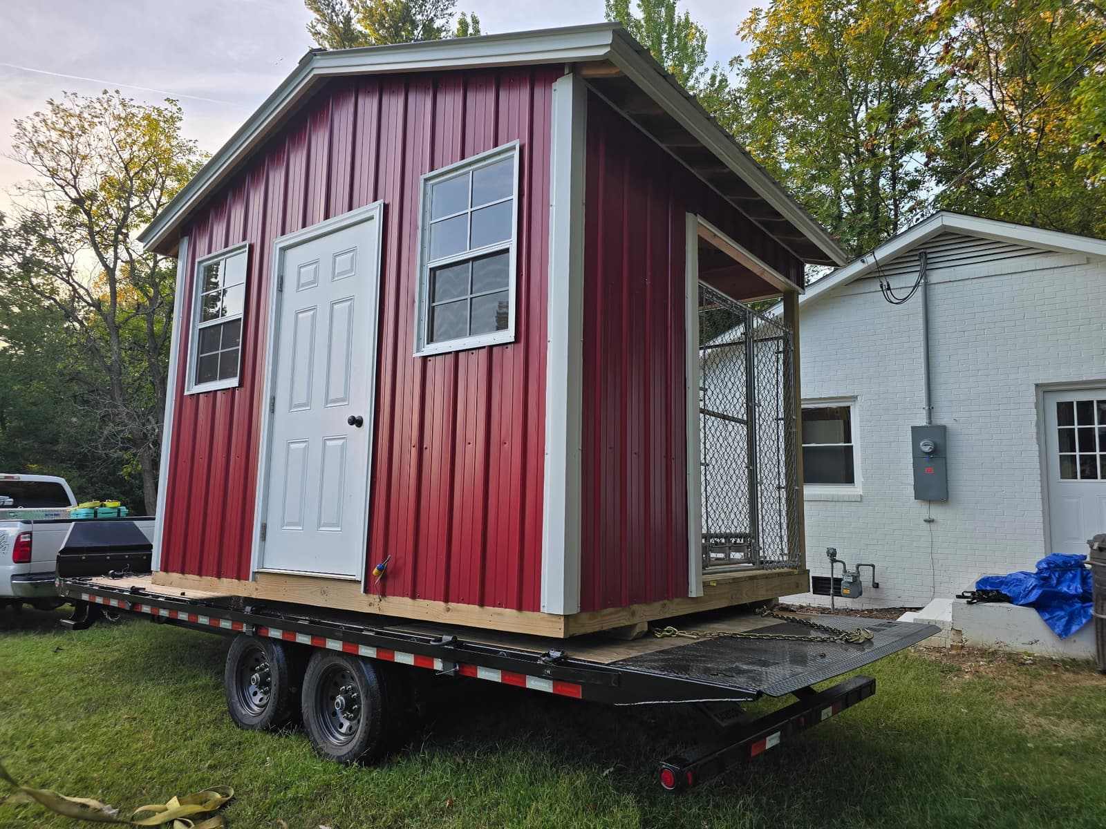 Close-up of a red barn-style shed with white trim, a white door, two windows, and an attached chain-link dog kennel, loaded on a black tandem-axle flatbed trailer