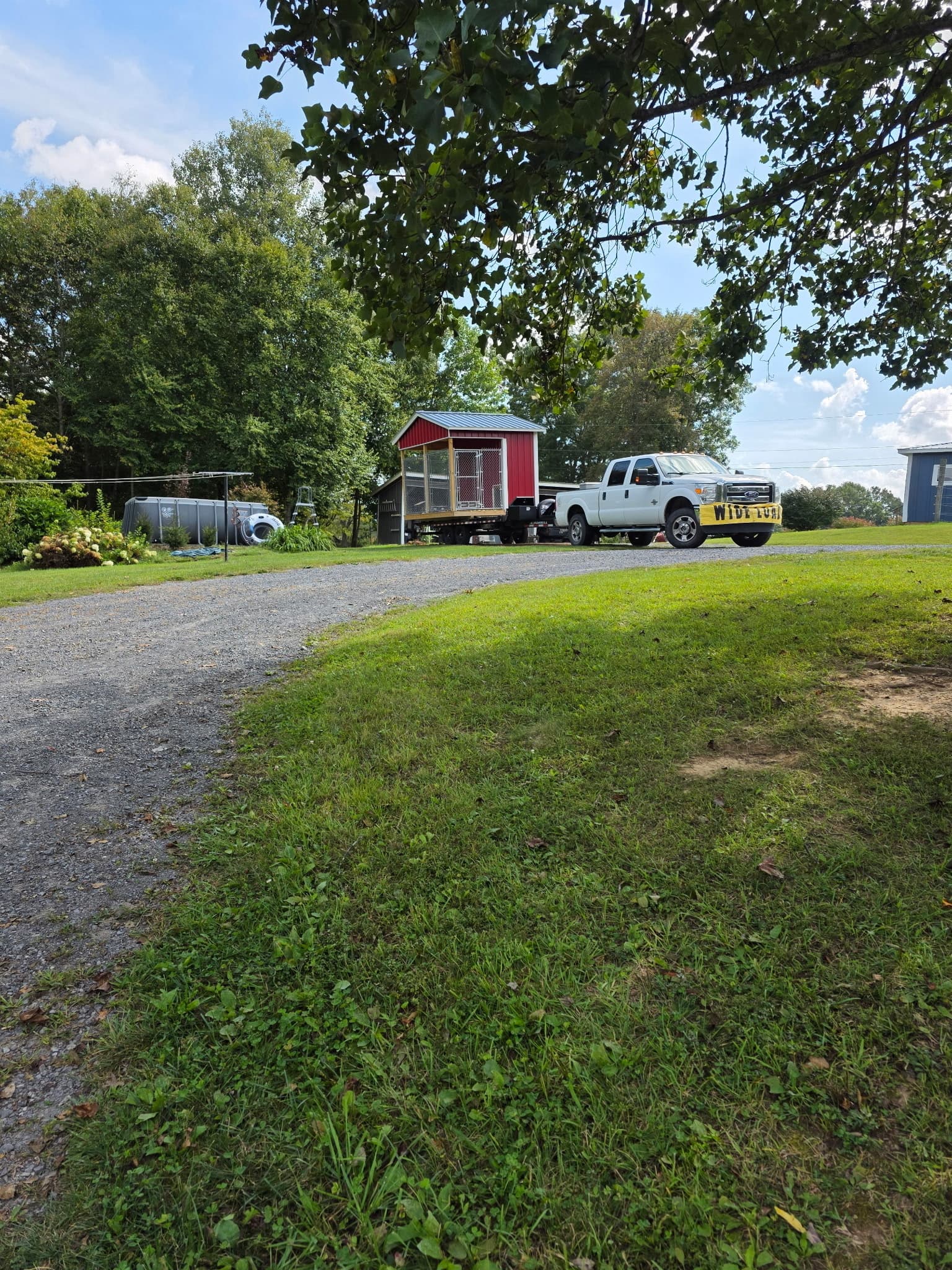 White Ford F-250 with a Wide Load banner towing a flatbed trailer loaded with a red barn-style shed down a gravel driveway surrounded by green lawn and trees