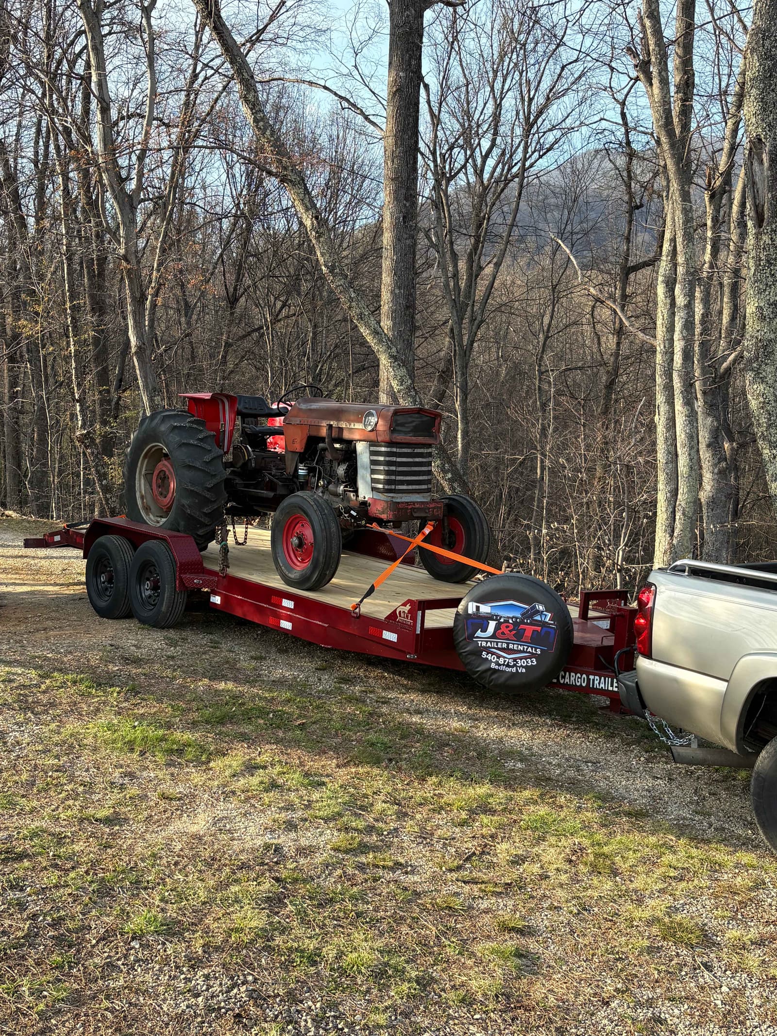 Vintage red farm tractor secured with orange tie-down straps on a J&T Trailer Rentals red dual-axle cargo trailer in a wooded rural setting