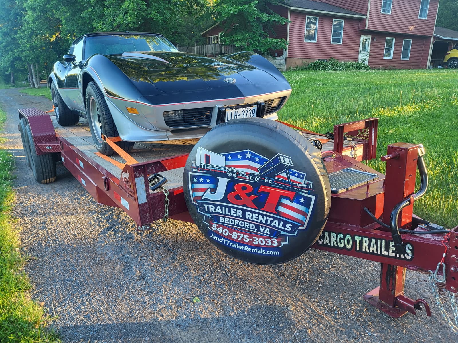 Black Corvette C3 loaded on the red J&T car trailer at dusk in a driveway with the branded spare tire cover prominently in the foreground
