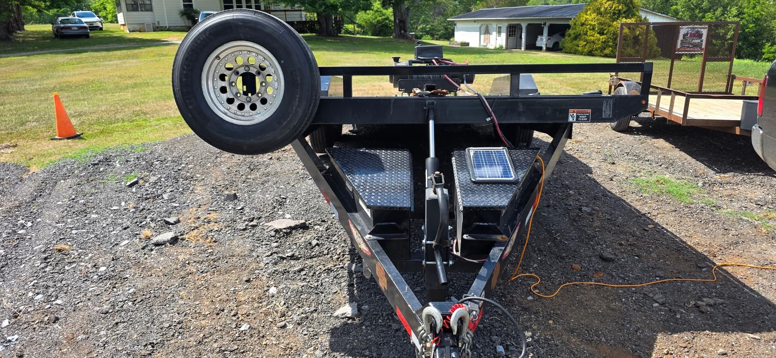 Front hitch end of the 24ft H&H tilt trailer showing the large spare tire, diamond plate toolbox, solar battery charger panel, winch cable and hydraulic tilt cylinder
