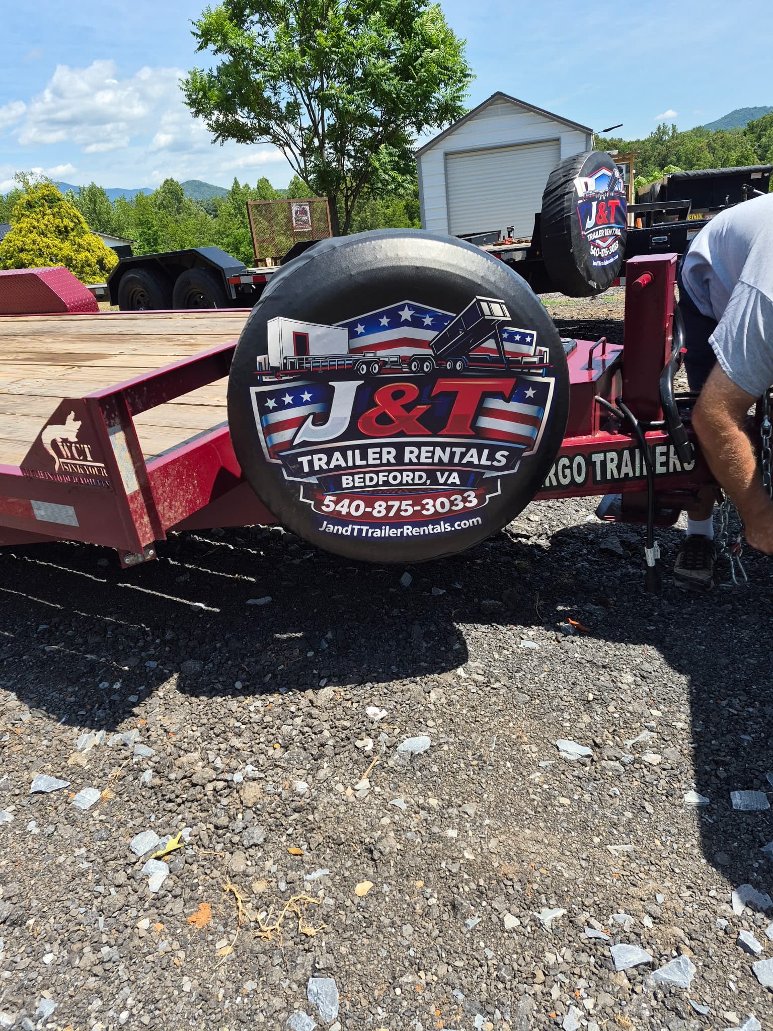 Close-up of the branded J&T Trailer Rentals spare tire cover showing the company logo, Bedford VA, 540-875-3033, and JandTTrailerRentals.com with two trailers visible in the background