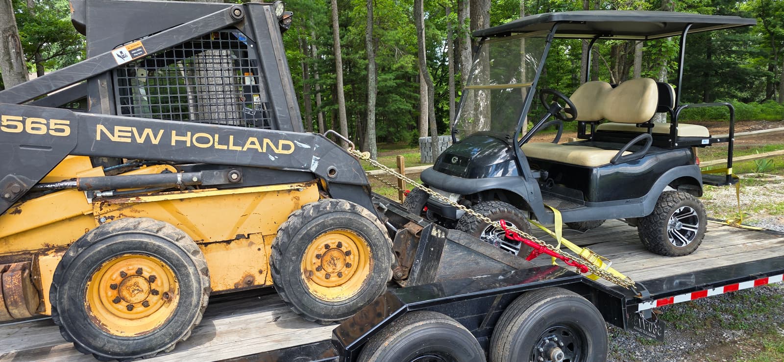 Close-up rear view of a New Holland L565 skid steer and a dark green golf cart chained side by side on the black 24ft tilt trailer wood deck