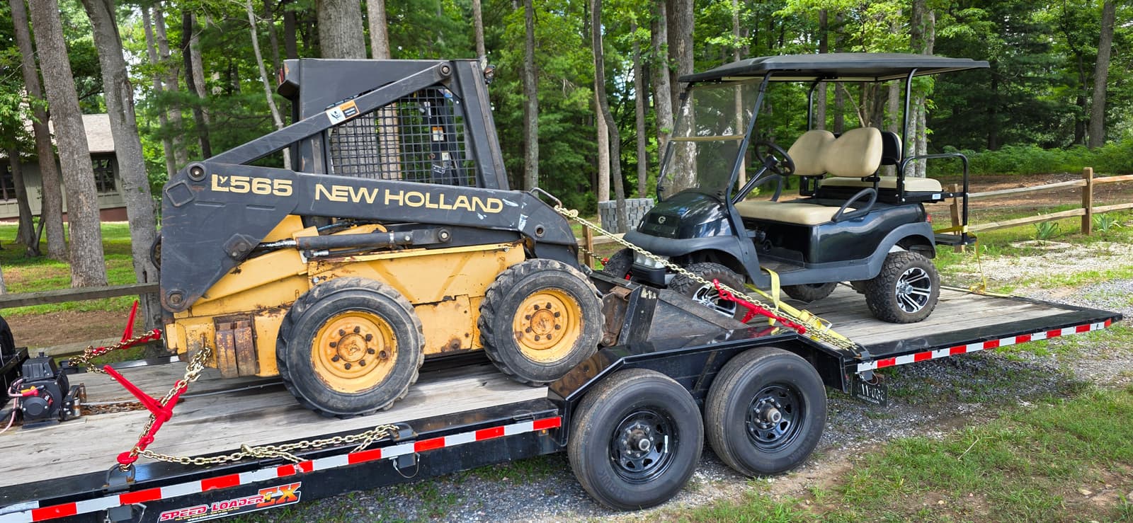 New Holland L565 skid steer loader and a dark green golf cart both loaded and chained on the black 24ft H&H tilt trailer, full side profile showing the wood deck and tandem axles