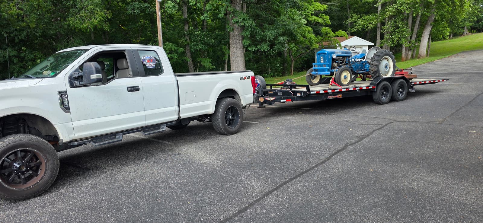 White lifted Ford F-250 4x4 with J&T Trailer Rentals sticker towing the black 24ft tilt trailer loaded with a classic blue Ford tractor on a paved road