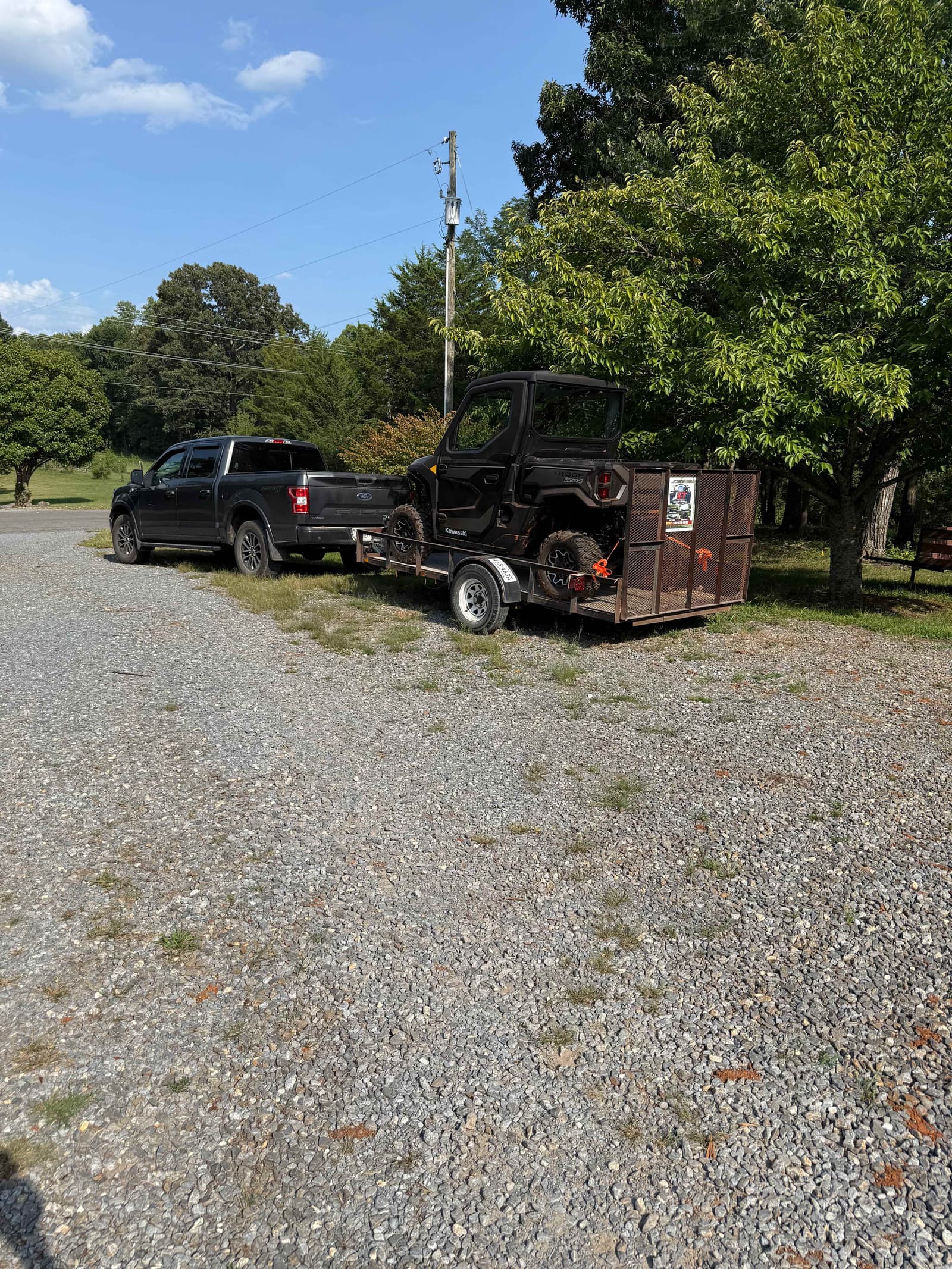 Kawasaki utility vehicle loaded on a trailer with a pickup truck and wooded property in the background