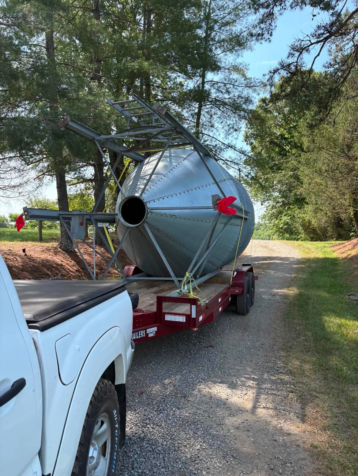 Large galvanized metal grain bin hopper cone secured with straps on a red flatbed gooseneck trailer with red safety flags, being towed down a tree-lined gravel driveway